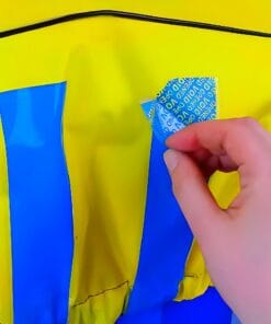 Ręka osoby odkładającej niebieską naklejkę z napisem na żółtej powierzchni, będącej częścią odzieży ochronnej. Obraz ilustruje aspekty związane z bezpieczeństwem pracy i ich znaczenie w ochronie środowiska.