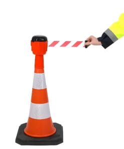 Pracownik w żółtej odzieży ochronnej rozciąga czerwono-białą taśmę ostrzegawczą z ustawionego stożka drogowego, co jest istotnym elementem zarządzania bezpieczeństwem pracy oraz ochroną środowiska w trakcie prac budowlanych lub awaryjnych.