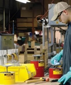 Mężczyzna w rękawicach ochronnych pracuje w warsztacie, porządkując narzędzia. Na stole widać czerwone pojemniki z ostrzeżeniem o niebezpieczeństwie oraz żółtą skrzynkę z elementami metalowymi. Dbałość o bezpieczeństwo pracy i środki awaryjne są kluczowe w tym środowisku.