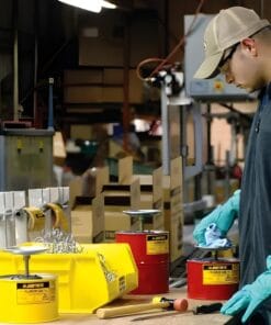 Mężczyzna w rękawicach ochronnych pracuje przy stole roboczym w warsztacie, obsługując różne narzędzia i pojemniki z substancjami chemicznymi. W tle widoczna jest zorganizowana przestrzeń robocza, a także środki awaryjne do zachowania bezpieczeństwa pracy i ochrony środowiska.