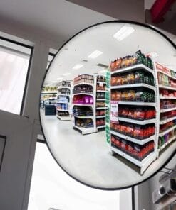 Wnętrze sklepu z widokiem na półki z różnorodnymi produktami, w tym przekąskami i artykułami gospodarczymi. Jasne oświetlenie i porządne rozmieszczenie towarów na białych regałach. Obok drzwi wejściowych widoczny odblask lusterka bezpieczeństwa, podkreślającego dbałość o bezpieczeństwo pracy w sklepie.