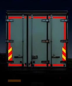 Tył ciężarówki z wyraźnymi, odblaskowymi oznaczeniami w kolorze czerwonym i żółtym, podkreślającymi bezpieczeństwo transportu. Ciemne tło sugeruje nocne warunki pracy, a solidna konstrukcja drzwi z zamkami świadczy o ochronie ładunku i przestrzeganiu norm bezpieczeństwa.