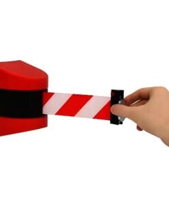 Zbliżenie na rękę odsuwającą taśmę ostrzegawczą o czerwono-białych paskach z urządzenia zamocowanego na ścianie, które służy do oznaczania strefy niebezpiecznej, promując bezpieczeństwo pracy.