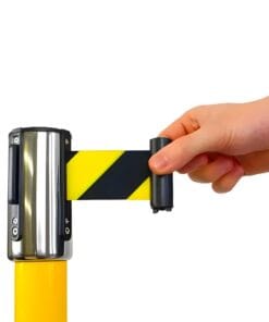 Ręka wysuwająca żółto-czarną taśmę ostrzegawczą z metalowego uchwytu. Elementy bezpieczeństwa pracy w środowisku przemysłowym, związane z ochroną środowiska i stosowanymi środkami awaryjnymi.