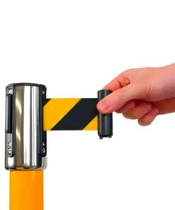 Ręka odciągająca taśmę ostrzegawczą w czarno-żółte paski z metalowego uchwytu. Element ten służy do oznaczania stref niebezpiecznych, zwiększając bezpieczeństwo pracy.