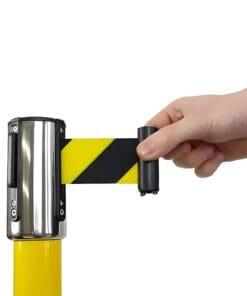 Zbliżenie ręki, która rozwija żółto-czarną taśmę ostrzegawczą z rolki zamocowanej na słupku, ilustrując zastosowanie zabezpieczeń w miejscu pracy dla ochrony środowiska i bezpieczeństwa.
