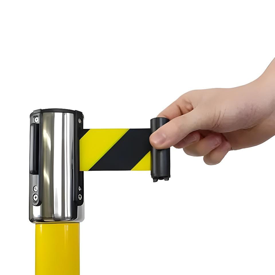 Zbliżenie ręki, która rozwija żółto-czarną taśmę ostrzegawczą z rolki zamocowanej na słupku, ilustrując zastosowanie zabezpieczeń w miejscu pracy dla ochrony środowiska i bezpieczeństwa.