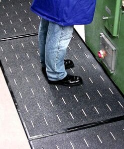 Osoba w niebieskim fartuchu i ciemnych butach stoi na antypoślizgowej powierzchni podłogi w zakładzie przemysłowym, z widocznymi oznaczeniami bezpieczeństwa. Materiał podłogowy zapewnia ochronę przed poślizgnięciem, co jest kluczowe dla bezpieczeństwa pracy w środowisku przemysłowym.