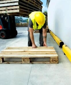 Pracownik w żółtej kamizelce i kasku położony na ziemi paletę drewnianą w miejscu pracy. W tle widać wózek widłowy oraz żółte elementy zabezpieczające. Scena ilustruje bezpieczeństwo pracy i organizację przestrzeni roboczej.