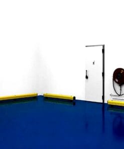 Widok wnętrza z niebieską podłogą oraz białymi ścianami. W rogu znajdują się żółte bariery ochronne, a obok wyjścia jest sprzęt awaryjny - wąż gaśniczy. Obraz ilustruje aspekty bezpieczeństwa pracy oraz środki awaryjne w zakładzie przemysłowym.