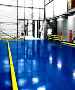 Jasna, błyszcząca podłoga w magazynie z żółtymi barierkami ochronnymi oraz wyjściami, idealne miejsce do zapewnienia bezpieczeństwa pracy oraz ochrony środowiska. Przestrzeń z widocznymi drzwiami i sprzętem przeciwpożarowym, co podkreśla znaczenie stosowania środków awaryjnych.