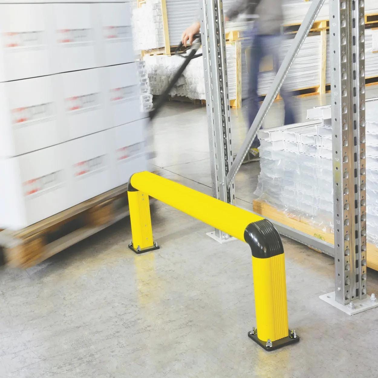 Żółta bariera ochronna w magazynie, chroniąca przed uderzeniami w regały. W tle widoczny pracownik przejeżdżający wózkiem widłowym z kartonami, co podkreśla znaczenie bezpieczeństwa pracy i organizacji przestrzeni w logistyce.