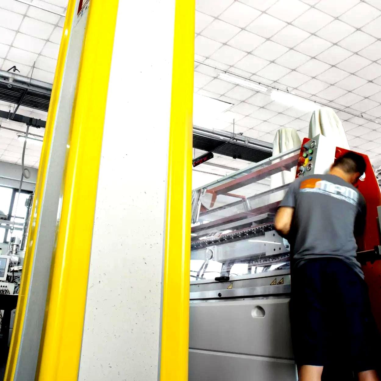 Zdjęcie przedstawia pracownika obsługującego nowoczesną maszynę do obróbki w przemyśle, z widocznymi elementami ochrony bezpieczeństwa na żółtych słupach. Materiały używane w produkcji podkreślają znaczenie bezpieczeństwa pracy i innowacji w technologii przemysłowej.