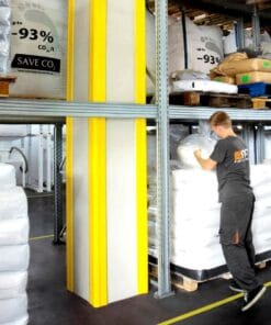 Mężczyzna pracujący w magazynie w otoczeniu worków z materiałami. W tle regały z opakowaniami, a pomiędzy nimi widoczny słup zabezpieczony żółtymi osłonami. Obraz ilustruje bezpieczeństwo pracy oraz właściwe przechowywanie materiałów, co jest istotne dla ochrony środowiska.