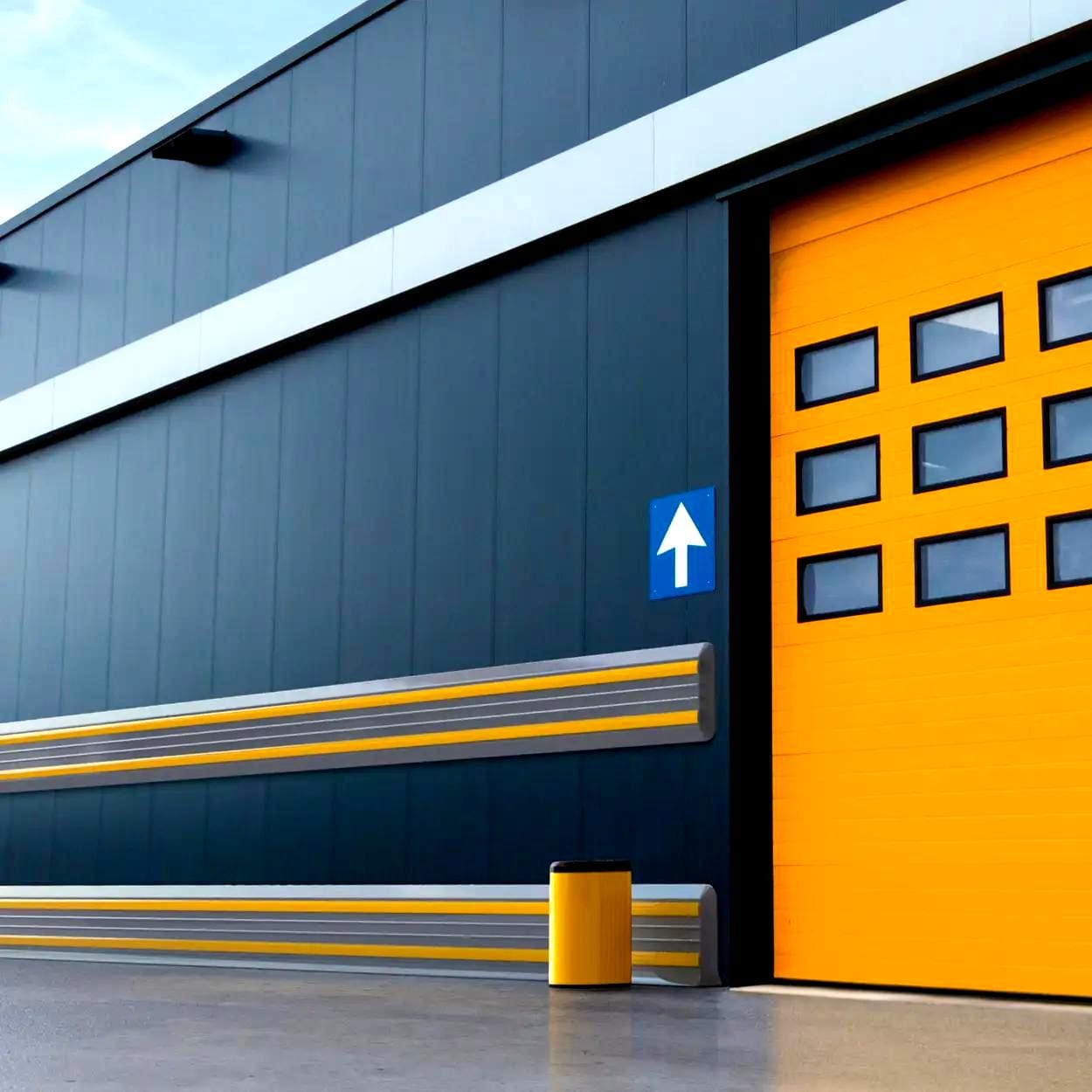 Zewnętrzna ściana nowoczesnego budynku przemysłowego z dużymi żółtymi drzwiami, oznaczeniem kierunkowym oraz elementami bezpieczeństwa, w tym zbiornikiem na odpady. Podkreśla dbałość o środowisko i bezpieczeństwo pracy, z funkcjonalnym i estetycznym designem.