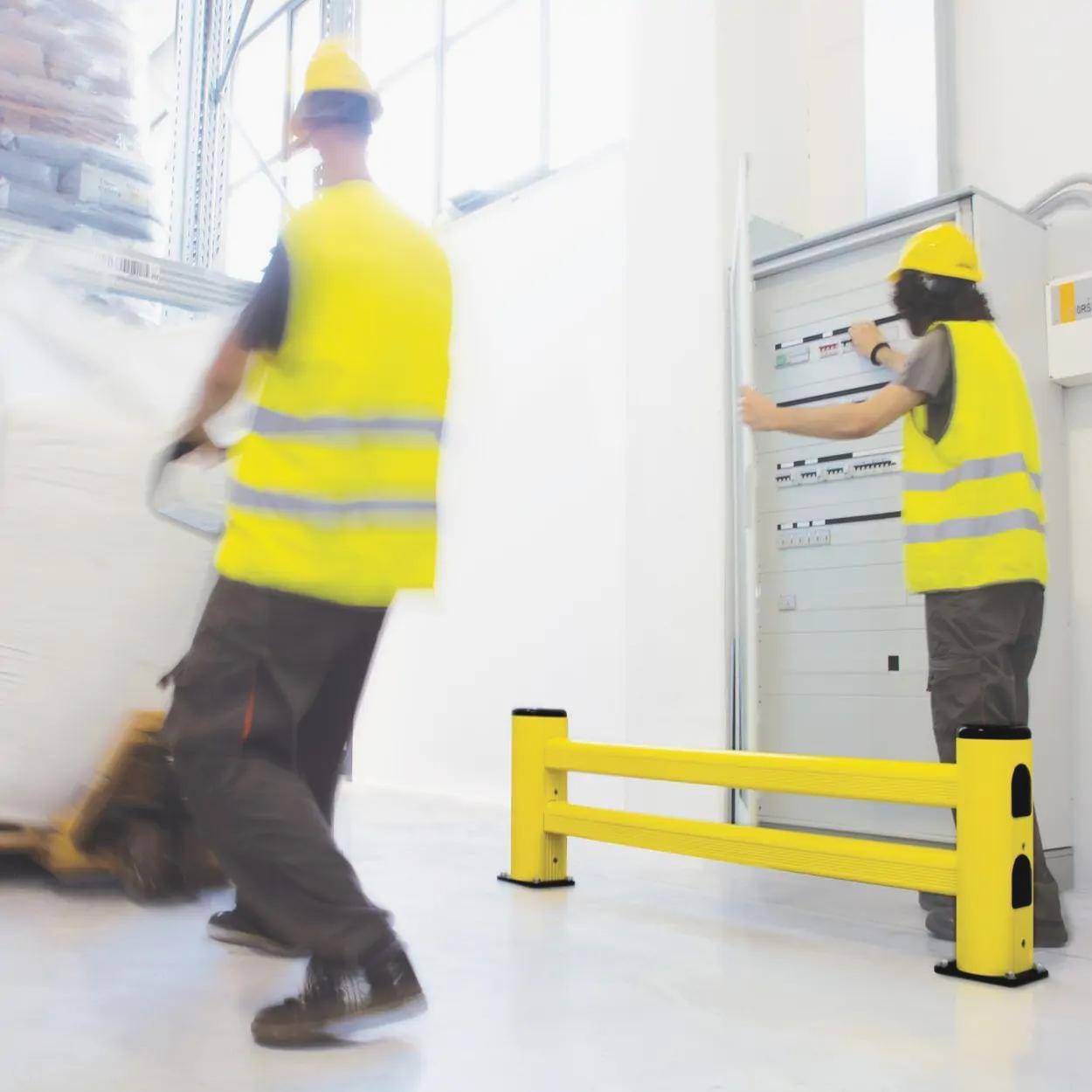 Pracownicy magazynu w odblaskowych kamizelkach żółtych, transportujący ładunek na wózku paletowym. W tle widoczny panel elektryczny oraz żółty element zabezpieczający, podkreślający istotę bezpieczeństwa pracy i ochrony środowiska w miejscu pracy.