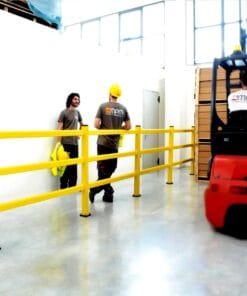 Mężczyźni w kaskach rozmawiają w pomieszczeniu z żółtym ogrodzeniem zabezpieczającym obszar roboczy. W tle widoczny wózek widłowy oraz sterty pudeł, co podkreśla znaczenie bezpieczeństwa pracy w środowisku magazynowym.