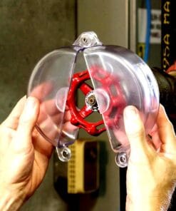 Obraz przedstawia osobę trzymającą przezroczysty osłonę, która odkrywa czerwony zawór. Akcesorium to może być element do monitorowania lub zabezpieczania systemów w kontekście ochrony środowiska lub bezpieczeństwa pracy.