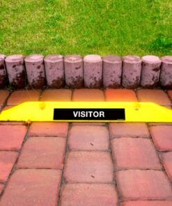 Żółty znak "VISITOR" umieszczony na kostce brukowej, wyróżniający się na tle zielonej trawy i kamiennego ogrodzenia. Elementy wizualne sugerują dbałość o bezpieczeństwo dostępu dla gości.