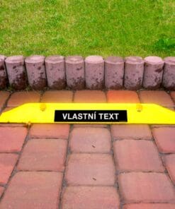Żółty znak z napisem „VLASTNÍ TEXT” na kostce brukowej, otoczony zieloną trawą i kamiennym obrzeżem, symbolizuje bezpieczeństwo i organizację przestrzeni. Dobrze widoczny element podkreślający znaczenie przestrzegania zasad.