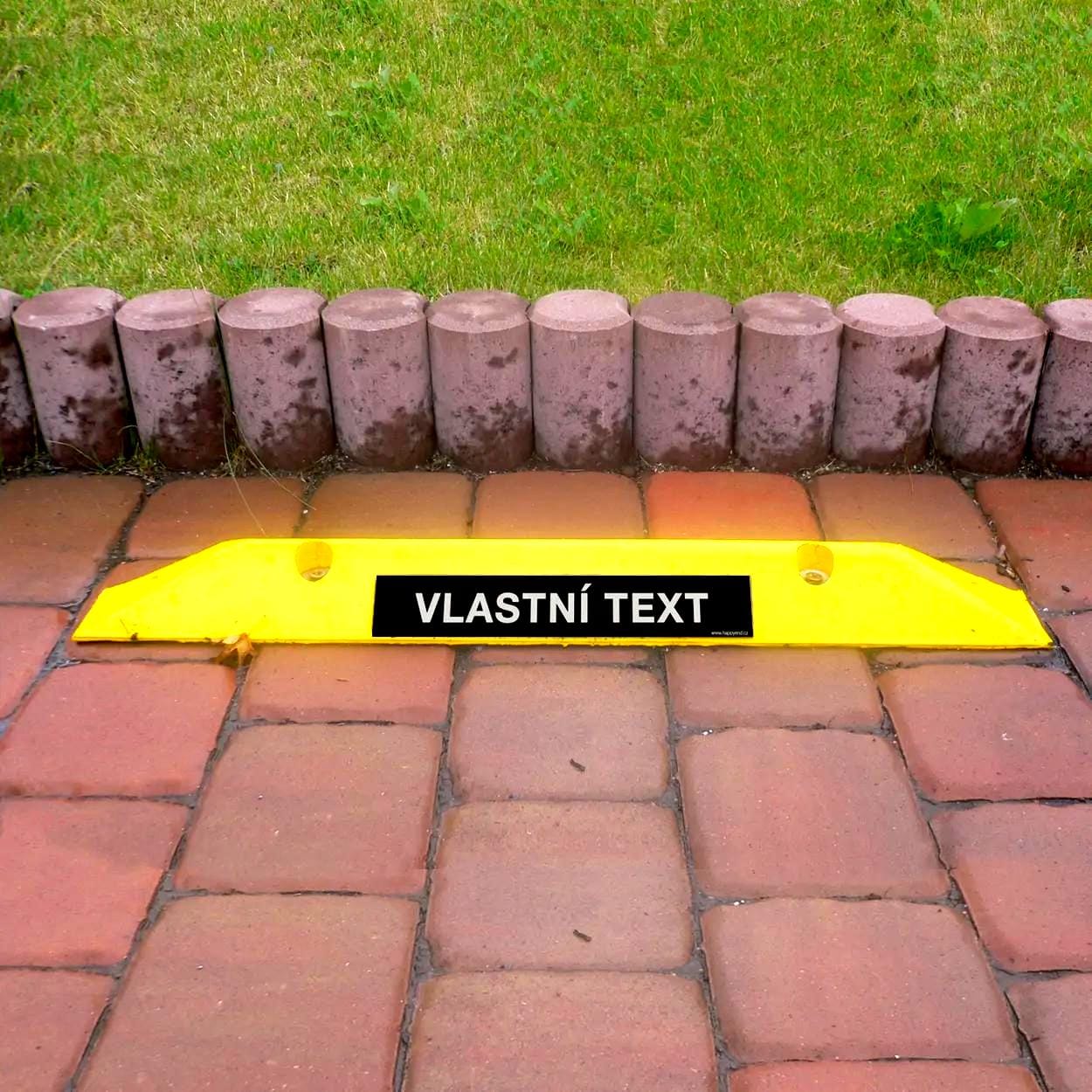 Żółty znak z napisem „VLASTNÍ TEXT” na kostce brukowej, otoczony zieloną trawą i kamiennym obrzeżem, symbolizuje bezpieczeństwo i organizację przestrzeni. Dobrze widoczny element podkreślający znaczenie przestrzegania zasad.