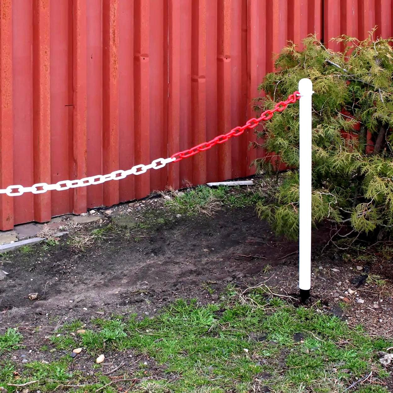 Biało-czerwony łańcuch zabezpieczający przymocowany do białego słupka, w tle widoczne czerwone kontenery i zieleń krzewu. Elementy te wskazują na działania związane z bezpieczeństwem i ochroną terenu, podkreślając znaczenie środków awaryjnych w kontekście ochrony środowiska.