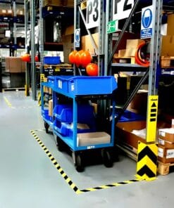Widok magazynu z niebieskimi wózkami transportowymi, poukładanymi kartonami oraz elementami zabezpieczeń. W tle widać regały z zapasami, a żółto-czarne oznaczenia na podłodze wskazują strefy bezpieczeństwa. Użyto środków awaryjnych dla zapewnienia bezpieczeństwa pracy.