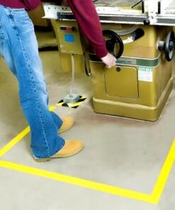 Pracownik obsługujący maszynę stolarską w bezpiecznej strefie oznaczonej żółtą taśmą izolacyjną. Zaznaczone miejsca awaryjne dla zachowania bezpieczeństwa pracy. Ekologiczne podejście do organizacji miejsca pracy w warsztacie.