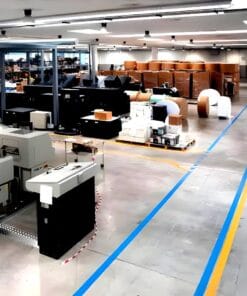 Przestronny magazyn z różnorodnym sprzętem i opakowaniami, wyposażony w nowoczesne urządzenia. Widoczne niebieskie i żółte linie na posadzce, co podkreśla zasady bezpieczeństwa pracy oraz organizację przestrzeni. Skoncentrowanie na efektywności i ochronie środowiska.