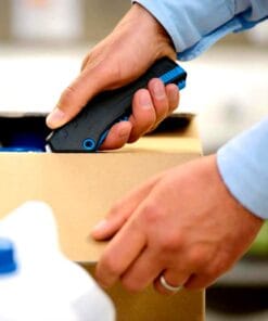 Osoba używająca noża do cięcia kartonu, trzymając go w prawej ręce. Praca z materiałami opakowaniowymi podkreśla znaczenie bezpieczeństwa pracy i odpowiednich praktyk w obszarze logistyki i magazynowania.