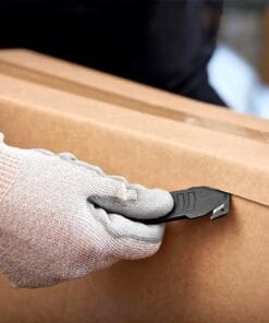 Ręka w rękawicy z materiału z narzędziem do otwierania kartonu, delikatnie przeciągająca po krawędzi zamkniętego pudełka, co ilustruje praktyki związane z bezpieczeństwem pracy i skuteczne zarządzanie opakowaniami w logistyce.