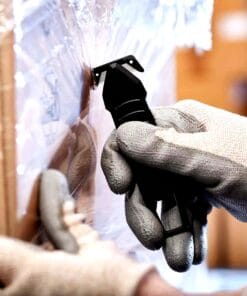 Osoba w rękawicach używa narzędzia do przecinania folii stretch na palecie. Obrazek ilustruje przygotowanie do transportu towarów, co jest ważne dla bezpieczeństwa pracy i efektywności operacji logistycznych.