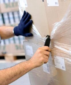 Mężczyzna w rękawicach otwiera paletę z kartonami, korzystając z noża. Praca w magazynie, bezpieczeństwo podczas rozpakowywania. Proces związany z obiegiem towarów i zarządzaniem zapasami. Ważne w kontekście ochrony środowiska i bezpieczeństwa pracy.