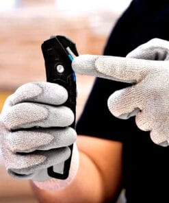 Osoba w rękawicach montażowych trzyma nóż z wymiennymi ostrzami, wskazując na mechanizm blokady. Narzędzie służy do cięcia, co podkreśla znaczenie bezpieczeństwa pracy podczas użycia ostrych przedmiotów.