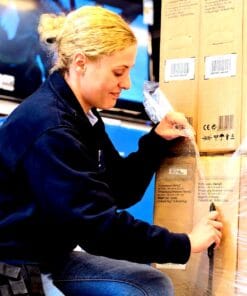 Kobieta rozpakowująca paczki z telewizorami w magazynie. Praca z towarem wymaga odpowiednich środków awaryjnych i bezpieczeństwa pracy. Podkreślenie ochrony środowiska poprzez efektywną logistykę.