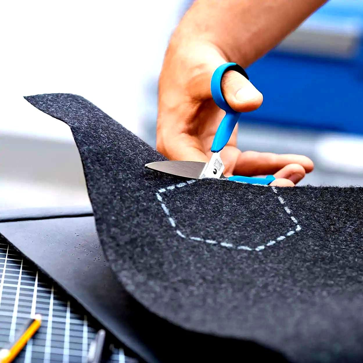 Osoba przycina czarny filc nożyczkami, wykonując precyzyjne cięcie zgodnie z zaznaczoną niebieską linią. W tle widać narzędzia oraz podłoże robocze, co ilustruje proces kreatywnej pracy. Idealne do tematyki ekologii i kreatywnych rozwiązań.