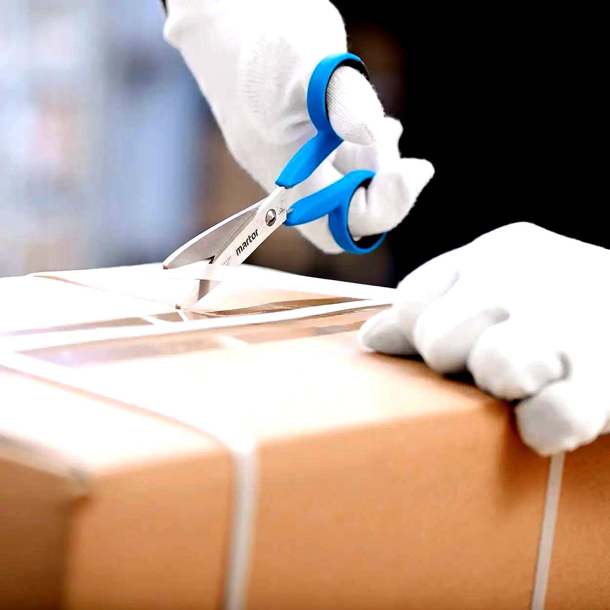 Osoba w rękawicach tnie taśmę na paczce za pomocą niebieskich nożyczek, co ilustruje proces otwierania przesyłek. Dbanie o środki awaryjne i bezpieczeństwo pracy w takich sytuacjach jest kluczowe dla ochrony środowiska i właściwego zarządzania materiałami.