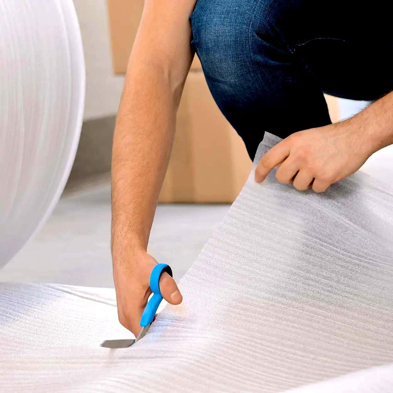 Mężczyzna przycinający folię bąbelkową nożyczkami, przygotowując materiał do pakowania. W tle widoczne kartony, co sugeruje działania związane z przeprowadzką lub transportem. Przykład zastosowania materiałów ochronnych w ekologicznym pakowaniu.