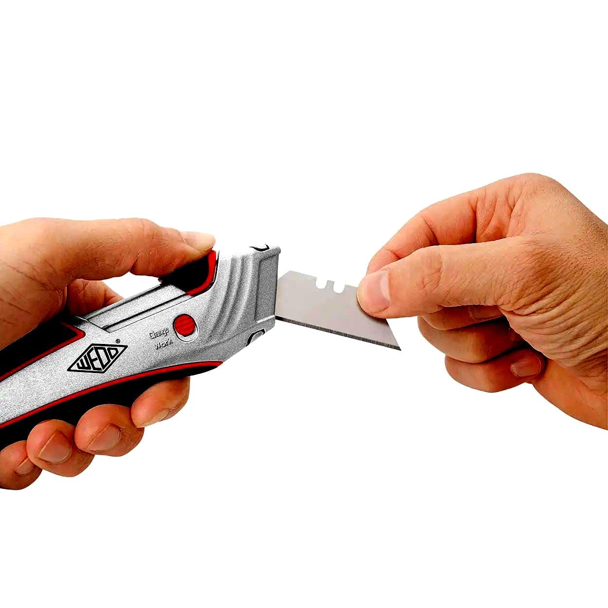 Osoba trzymająca nożyk z wymiennym ostrzem, demonstrująca sposób wymiany ostrza na urządzeniu. Narzędzie ma metaliczne wykończenie i jest wyposażone w funkcjonalne elementy, co podkreśla jego zastosowanie w przemyśle. Bezpieczeństwo pracy i efektywność są kluczowe w używaniu takich narzędzi.