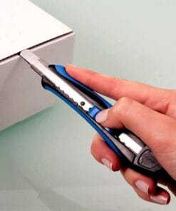 Osoba używająca noża do cięcia, aby otworzyć kartonowy pudełko. Narzędzie posiada niebieską i czarną rączkę, co wskazuje na bezpieczeństwo pracy z ostrym narzędziem. Przykład efektywnego wykorzystania środków awaryjnych w codziennych czynnościach.