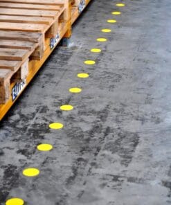 W magazynie widoczna jest linia żółtych znaczników na podłodze wskazujących bezpieczną ścieżkę, z drewnianymi paletami w tle. Elementy te są istotne dla bezpieczeństwa pracy i organizacji przestrzeni.