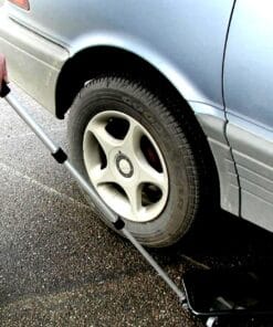 Mężczyzna używa narzędzia do sprawdzania ciśnienia w oponie samochodu. Opona jest zamontowana na srebrnym pojeździe, stojącym na szarym asfalcie. W tle widać fragment nadwozia auta. To ważny element bezpieczeństwa pracy oraz utrzymania pojazdu w dobrym stanie.