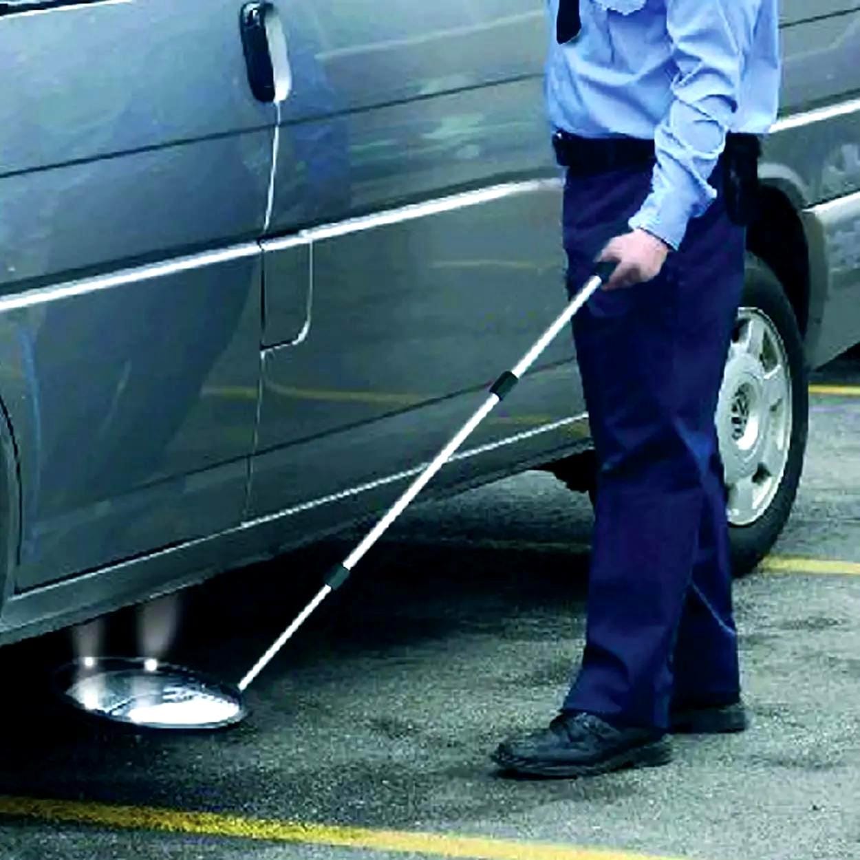 Mężczyzna w mundurze korzysta z długiego narzędzia, aby zbadać przestrzeń pod pojazdem. Obraz ilustruje działania związane z bezpieczeństwem pracy oraz monitorowaniem potencjalnych zagrożeń w obszarze parkingu, co jest istotne w kontekście ochrony środowiska i skutecznych środków awaryjnych.