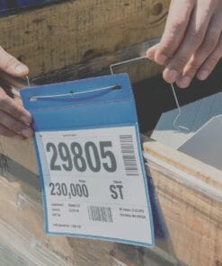 Zdjęcie przedstawia osobę trzymającą niebieską etykietę z numerem 29805, zawierającą szczegóły dotyczące magazynowania, takie jak waga i ilość. Etykieta jest mocowana do drewnianej skrzyni, co podkreśla znaczenie organizacji i bezpieczeństwa pracy w kontekście logistyki.