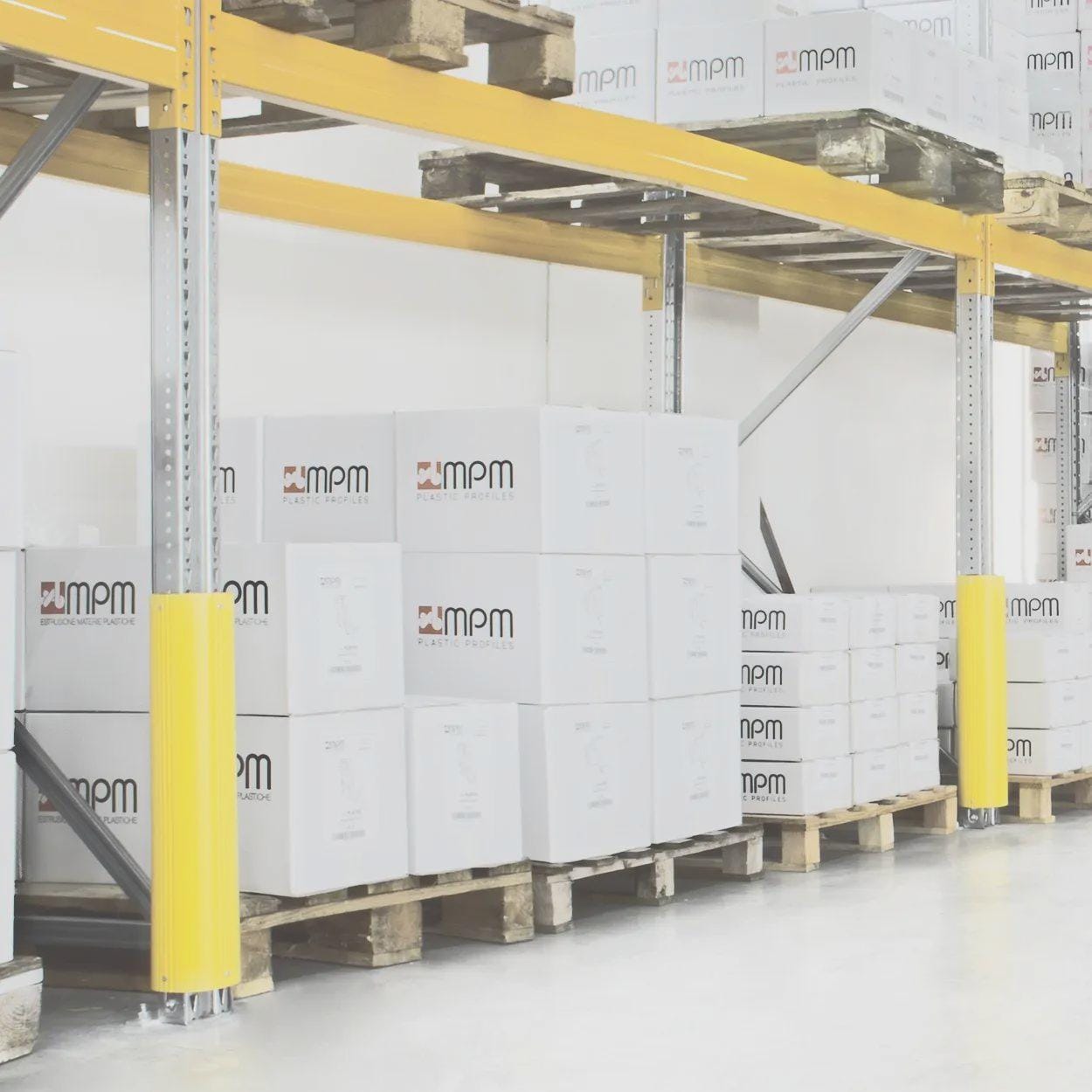 Zdjęcie przedstawia magazyn z regałami i paletami wypełnionymi białymi kartonami z logo MPM. Zastosowane środki awaryjne, takie jak żółte osłony, zapewniają bezpieczeństwo pracy i organizację przestrzeni w kontekście ochrony środowiska.