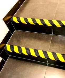 Zbliżenie na schody z żółto-czarnym oznaczeniem bezpieczeństwa, wskazującym na niebezpieczne krawędzie. Zastosowanie tych oznaczeń podnosi bezpieczeństwo w miejscu pracy, minimalizując ryzyko upadków. Schody wyłożone ciemnymi płytkami, z widocznymi detalami krawędzi.