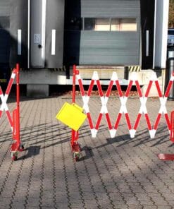 Złożone czerwono-białe barierki zabezpieczające, z żółtą tablicą, ustawione przed zamkniętymi drzwiami w hali. Elementy te służą do zapewnienia bezpieczeństwa pracy i oznaczenia obszaru zagrożonego w sytuacjach awaryjnych.