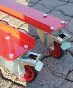 Zbliżenie na dwa czerwone kółka wózka przemysłowego, osadzone na stalowych szynach. Widać szczegóły konstrukcji, w tym nakrętki i śruby, a także fakturę podłoża. Elementy te są istotne dla bezpieczeństwa pracy i ochrony środowiska w zakładach przemysłowych.