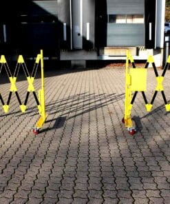 Żółty składany barierka z rolkami, umieszczona na kostkowanej powierzchni, zapewniająca bezpieczeństwo w obszarze prac budowlanych. Bariera może być używana do ochrony przed wjazdem w strefy ograniczone, co podkreśla znaczenie środków awaryjnych w zapewnieniu bezpieczeństwa pracy.