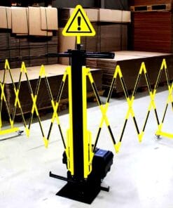 Przenośna bariera ostrzegawcza w kolorze żółtym i czarnym, z oznaczeniem ostrzeżenia, ustawiona w magazynie z kartonami. Element ten zapewnia bezpieczeństwo pracy oraz kontrolę dostępu w obszarze zagrożonym. Idealne rozwiązanie do zastosowania w środowiskach przemysłowych.